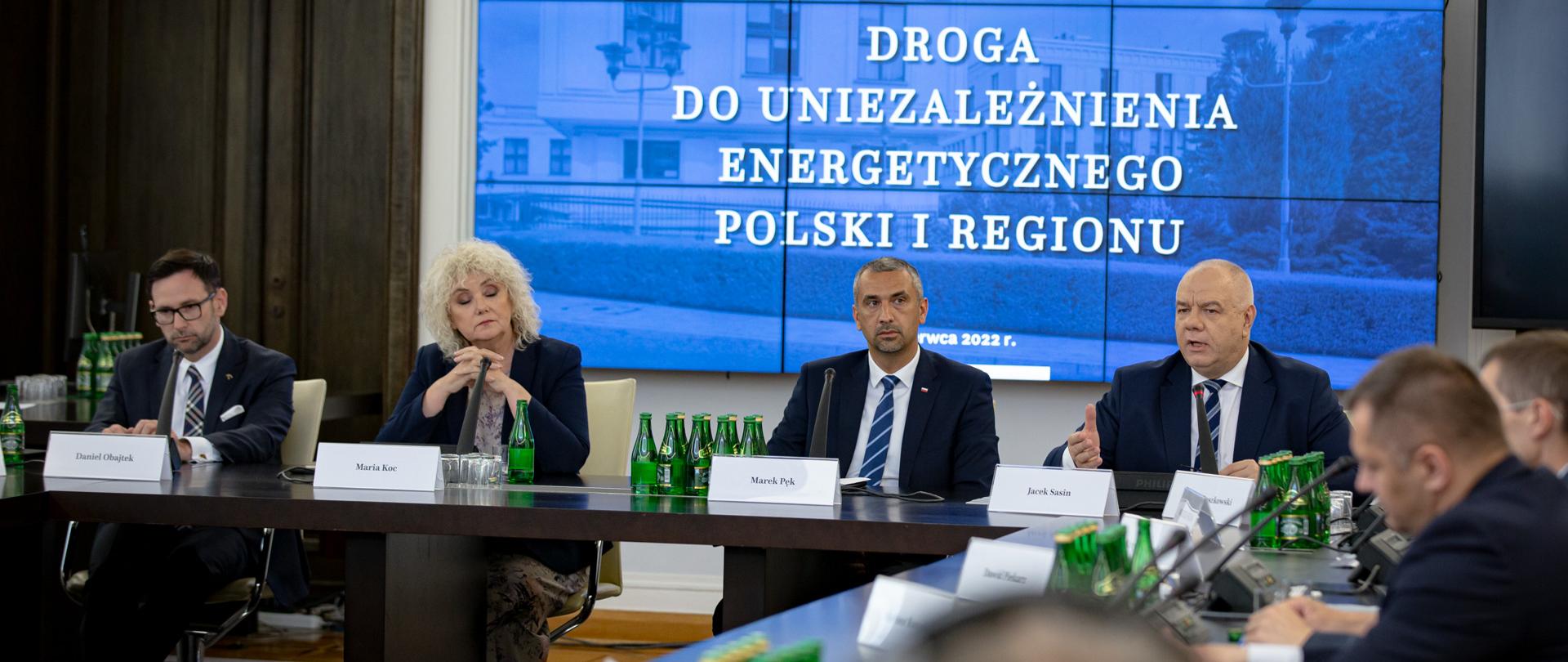 Wicepremier wraz z innymi uczestnikami konferencji siedzą za stołem. W tle ekran z napisem droga do uniezależnienia energetycznego Polski i regionu.
