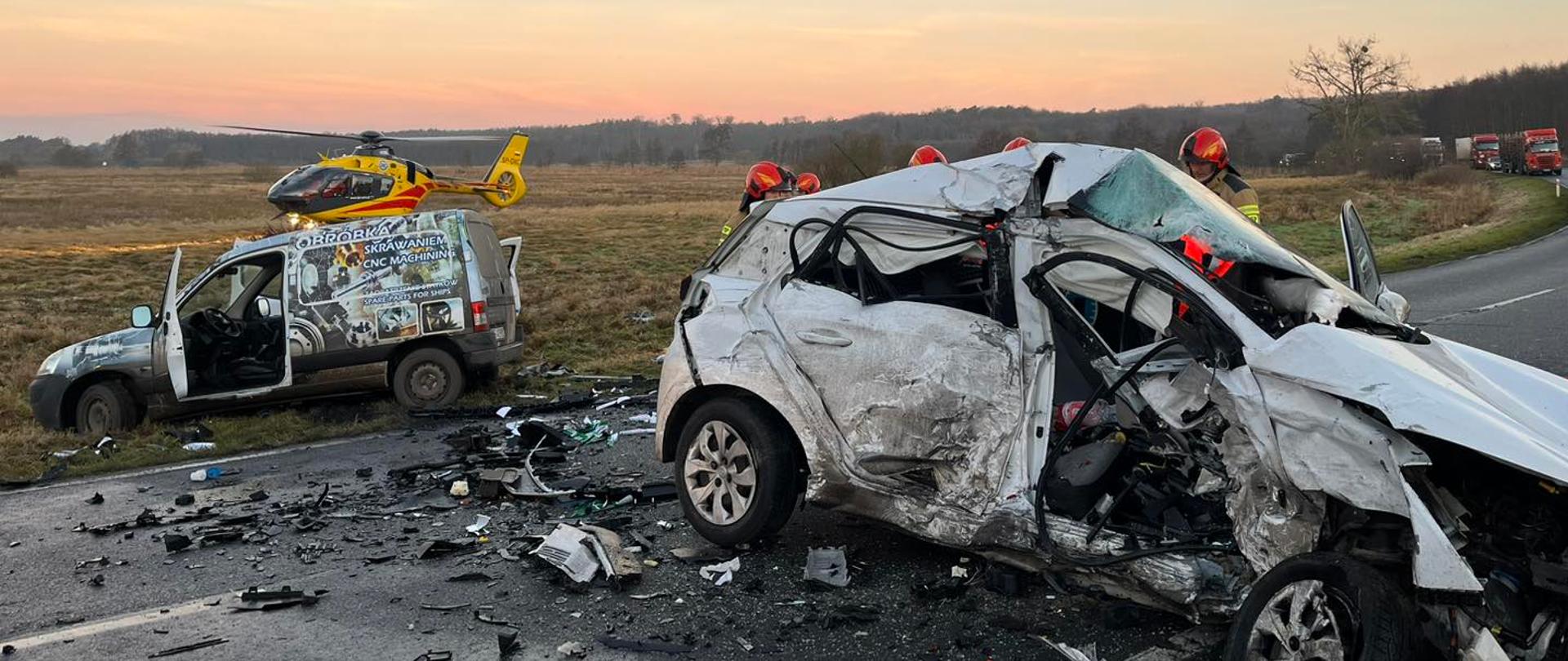 Na pierwszym planie zniszczony pojazd osobowy stojacy na drodze asfaltowej. Za nim strażacy w ubraniach specjalnych. Wkoło pojazdu pełno połamanych plastików i potłuczonego szkła. W tle dwa inne zniszczone pojazdy, które znajdują się poza jezdnią na poboczu. Za nimi na trawie stoi helikopter LPR.