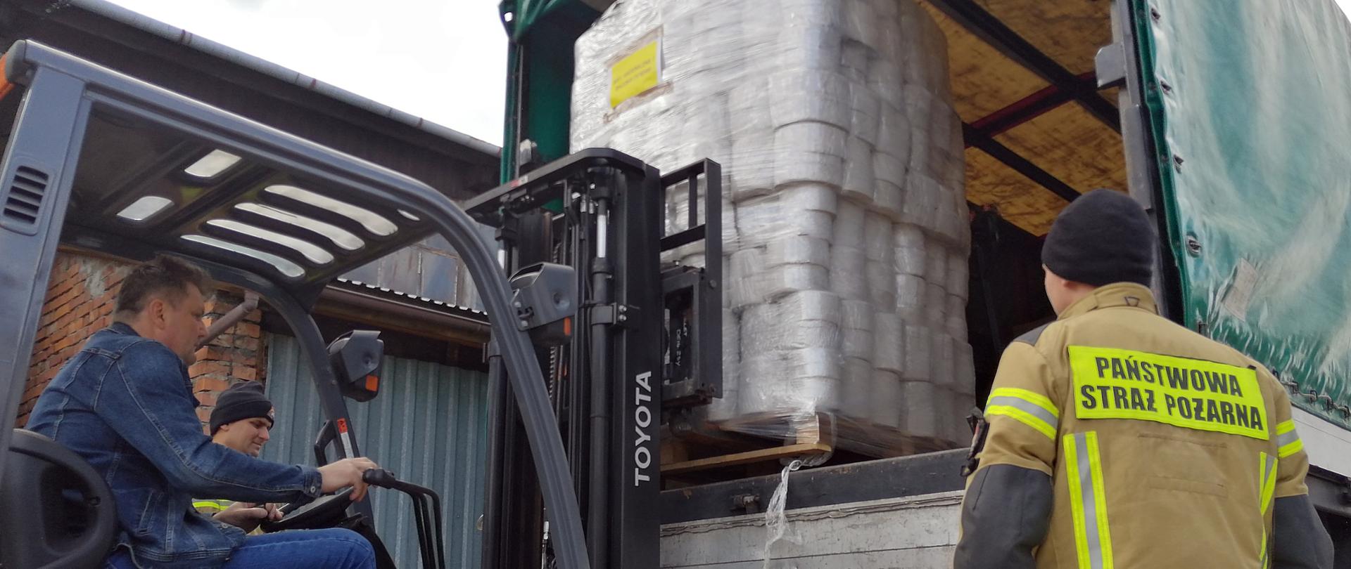 Załadunek artykułów higienicznych przy pomocy wózka widłowego na samochód ciężarowy. Po prawej stronie widoczny z tyłu strażak KP PSP w Wadowicach.