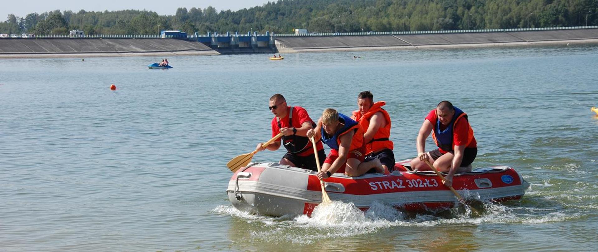 Na zdjęciu widać na akwenie wodnym rywalizację w wyścigach na wodzie łodziami wiosłowymi. Na pierwszym planie 4 strażaków ubranych w kamizelki ratownicze wiosłuję z pontonu.