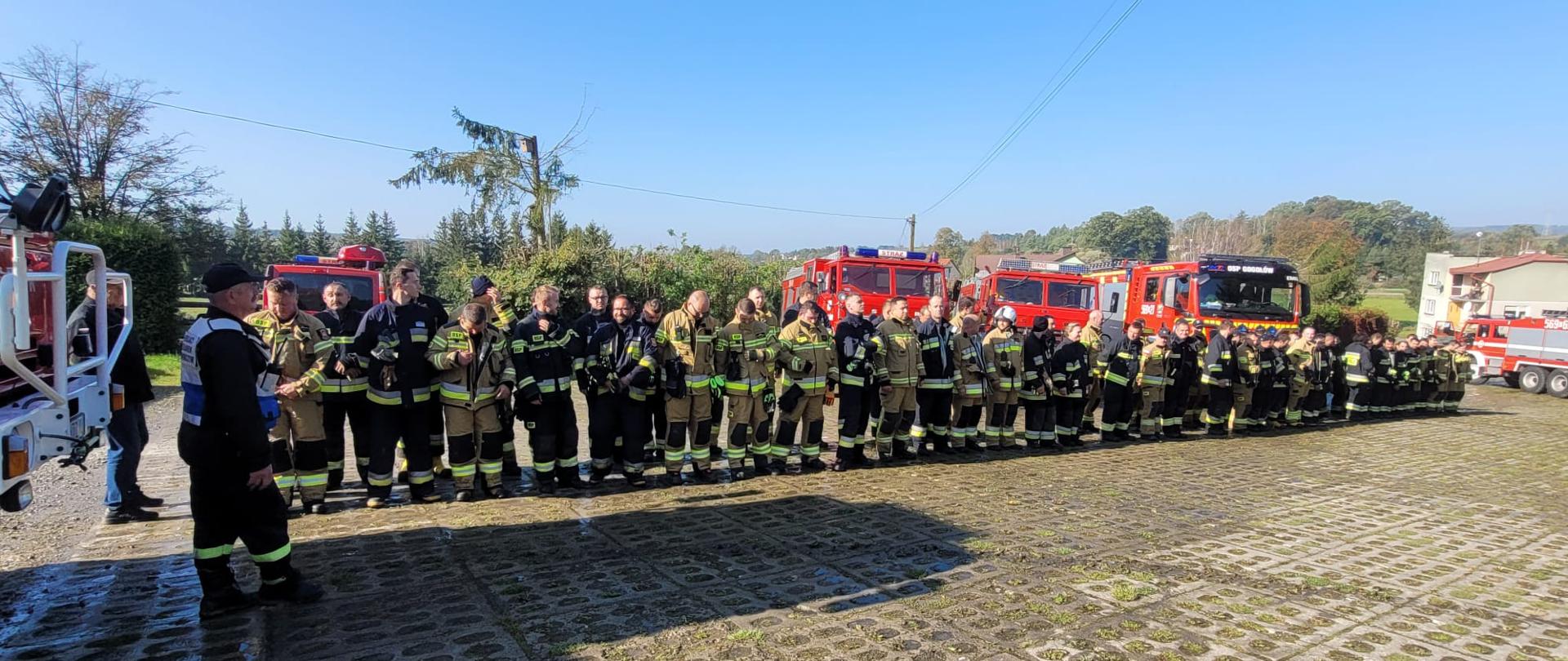 Strażaków ustawionych w dwuszeregu na tle samochodów pożarniczych, po prawej stronie stoi dowódca ubrany w kamizelkę w kolorze biało niebieskim.