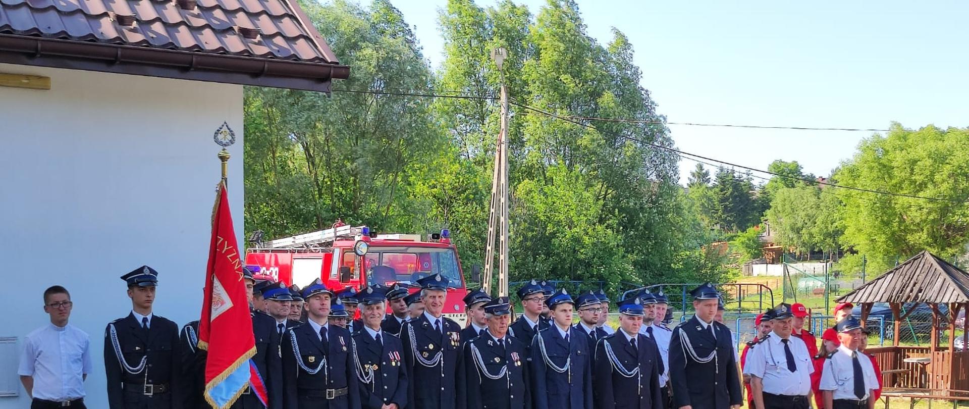 Na kostce brukowej stoją strażacy ubrani w galowe mundury i czapki na głowach. Trzech strażaków ma ubrane w białe rękawiczki. Jeden ze strażaków trzyma w ręku sztandar w kolorze czerwonym. Po prawie stronie stoją osoby ubrane w czerwone stroje i czapki z Młodzieżowych Drużyn Pożarniczych. Za strażakami stoi biały budynek z brązowym dachem. Po prawej stronie za strażakami stoi czerwony samochód pożarniczy a obok znajduje się drewniana altanka. Za samochodem pożarniczym jest słup energetyczny oraz drzewa.
