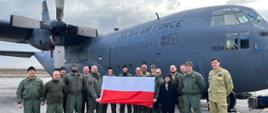 Grupa strażaków, wojskowych pilotów oraz obywatel Turcji na grupowym zdjęciu przed samolotem wojskowym. W ręku trzymają flagę polski
