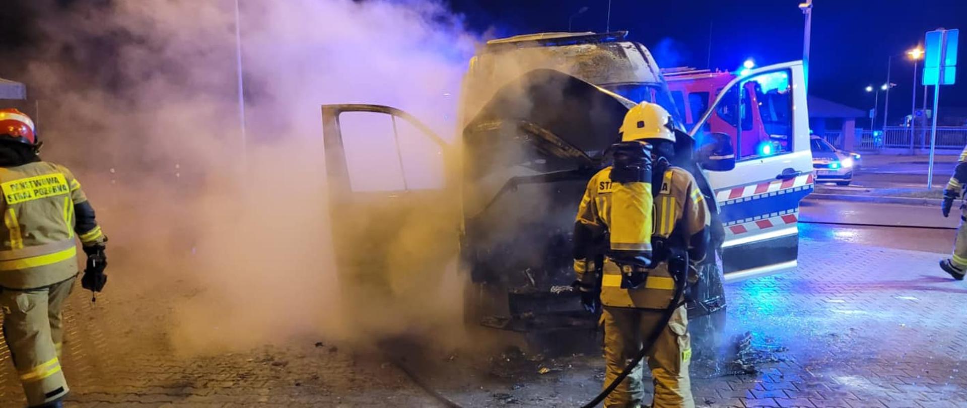 Na zdjęciu znajduje się palący się samochód oraz strażak gaszący pojazd