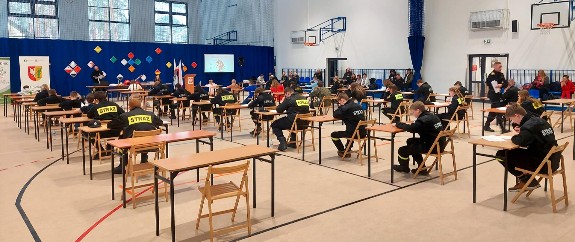 Na zdjęciu młodzież biorąca udział w Powiatowym Finale Ogólnopolskiego Turnieju Wiedzy Pożarniczej 2023. Zdjęcie na sali gimnastycznej.