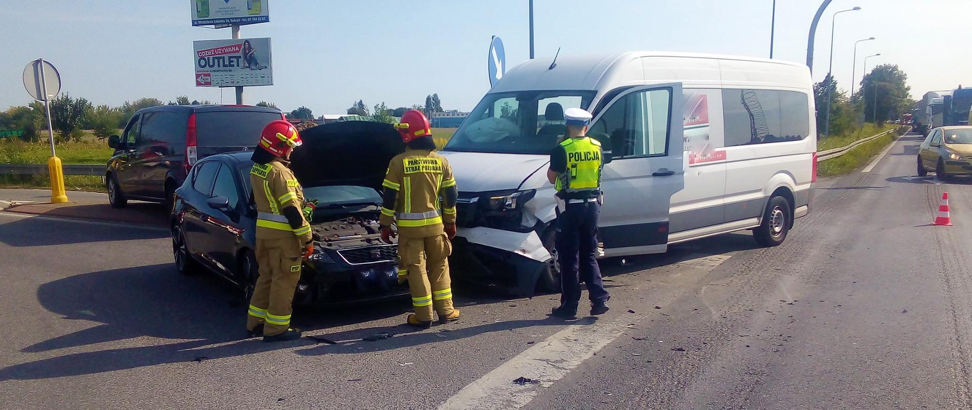 Zdjęcie przedstawia strażaków i policjanta działających przy wypadku drogowym.
W tle pojazdy uszkodzone, ulica i pola.
