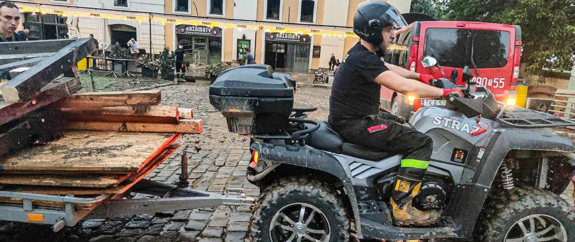 Strażak prowadzący quada, który ciągnie przyczepkę z drewnianymi belkami