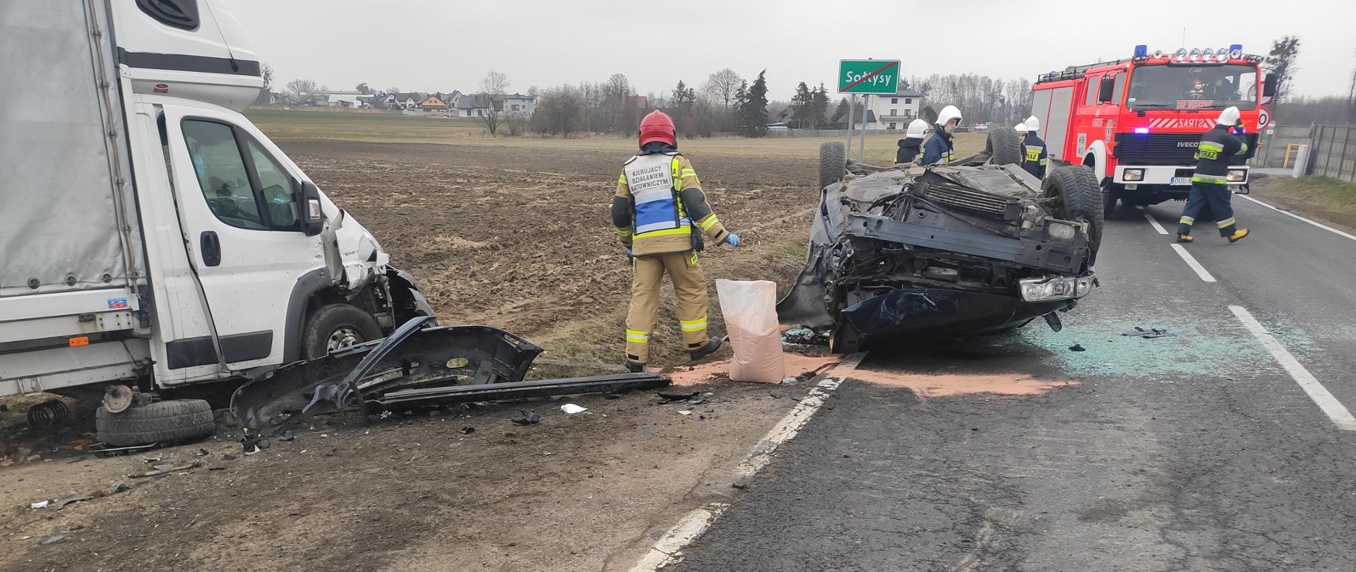 Zdjęcie przedstawia przewrócony na dachu samochód osobowy, po lewej stronie na polu stoi samochód dostawczy. Na zdjęciu widoczni również strażacy w ubraniach specjalnych koloru piaskowego i czarnego na głowie hełmy czerwone i białe. Z tył na drodze stoi samochód pożarniczy koloru czerwonego. Na drodze rozsypane sorbent, szkło oraz części karoserii samochodowej.