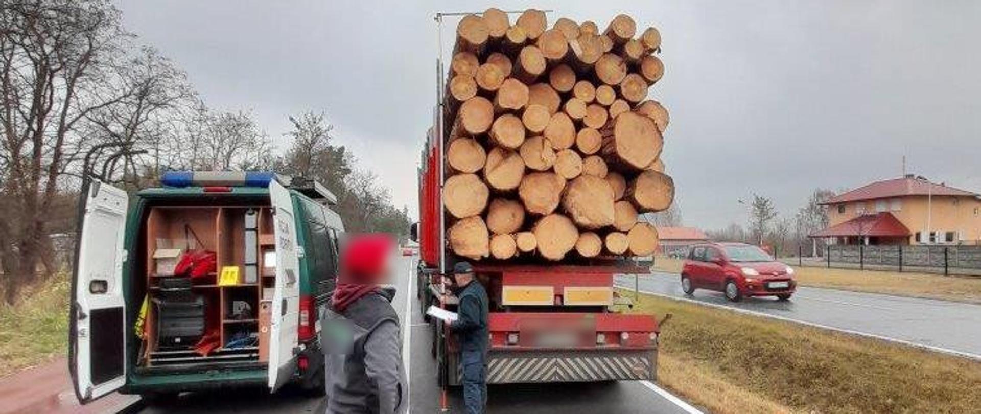 Inspektor z przymiarem stoi obok naczepy z kłodami drewna. Pomiarom przygląda się kierowca. Obok stoi radiowóz ITD.