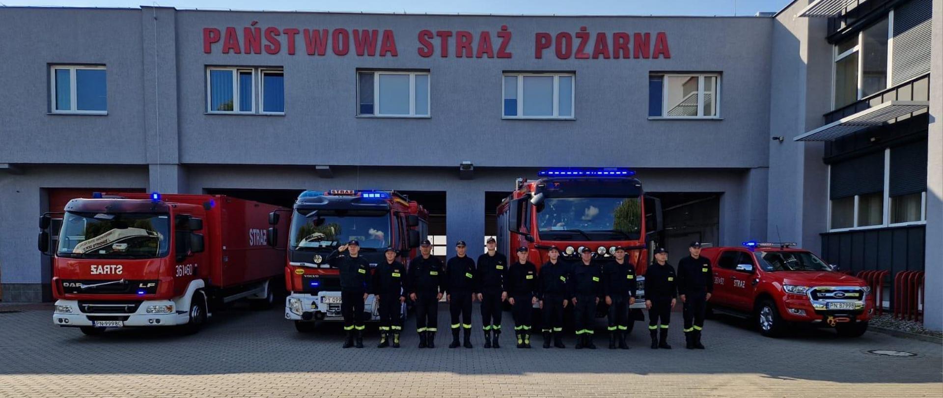 Zdjęcie przedstawia strażaków stojących w szyku przed garażem podczas oddania hołdu bohaterom powstania warszawskiego. Za nimi stoją samochody strażackie. 