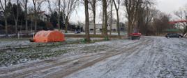 Na zdjęciu widać pomarańczowy namiot strażacki. Miejsce nad rzeką. Ośnieżone. W tle drzewa. Zachmurzenie lekkie. Między pojazdami strażacy W tle most.