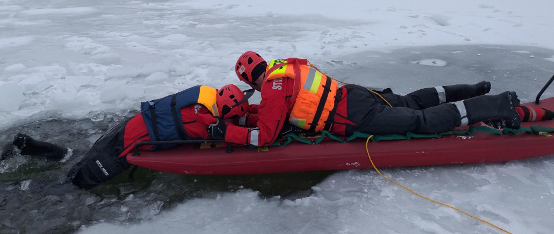 Zdjęcie przedstawia dwóch strażaków w ubraniach wypornościowych oraz kamizelkach asekuracyjnych. Mężczyźnie mają założone na głowach kaski ochronne. Znajdują się oni na środku zalewu miejskiego w Jędrzejowie, który pokryty jest lodem. Jeden ze strażaków, leżący na saniach lodowych wyciąga drugiego z przerębla. Na drugim planie widnieją budynki i bloki mieszkalne.
