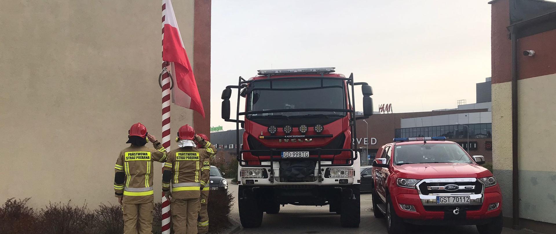 Zdjęcie przedstawia uroczyste podniesienie flagi, poczet flagowy stoi tyłem, po prawej stronie samochody strażackie, flaga państwowa wciągana jest na maszt