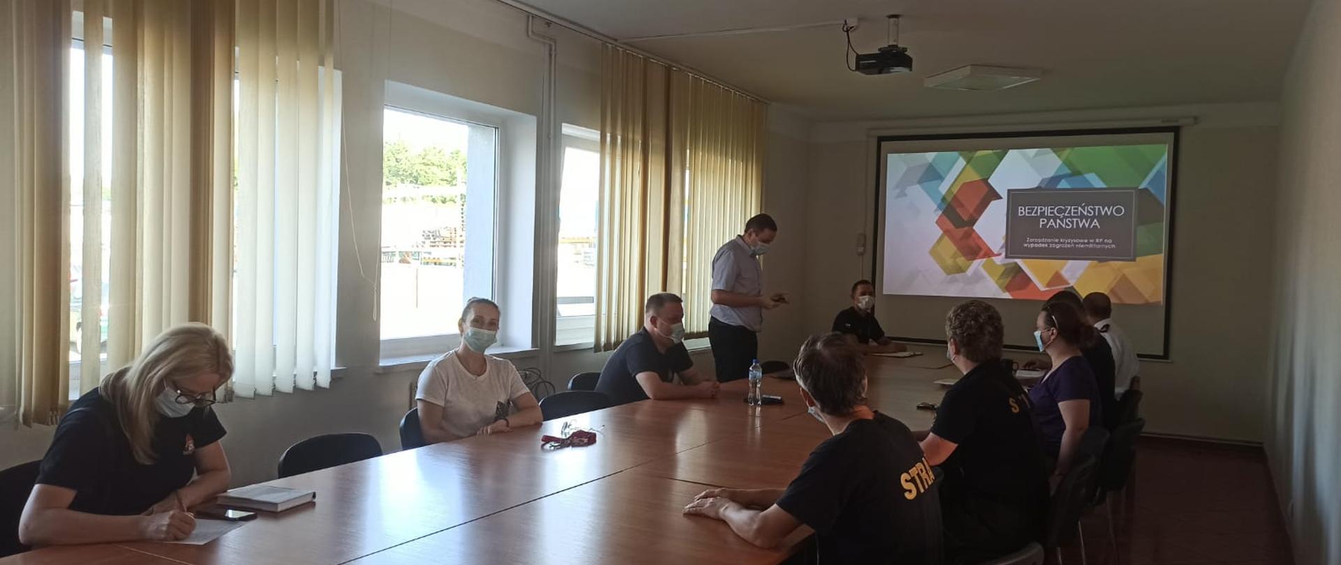 Zdjęcie przedstawia szkolenie, które odbywa się w sali szkoleniowej. W centrum zdjęcia widać stoły przy których siedzą funkcjonariusze uczestniczący w szkoleniu ubrani w czarne mundury oraz maseczki chirurgiczne. Jena osoba prowadząca szkolenie, która siedzi przy komputerze jest ubrana w niebieską służbową koszulę. W tle zdjęcia widać: drzwi, lampy alarmowe, rzutnik, tablice projekcyjną na której wyświetlony jest program do łączności, żaluzje okienne.