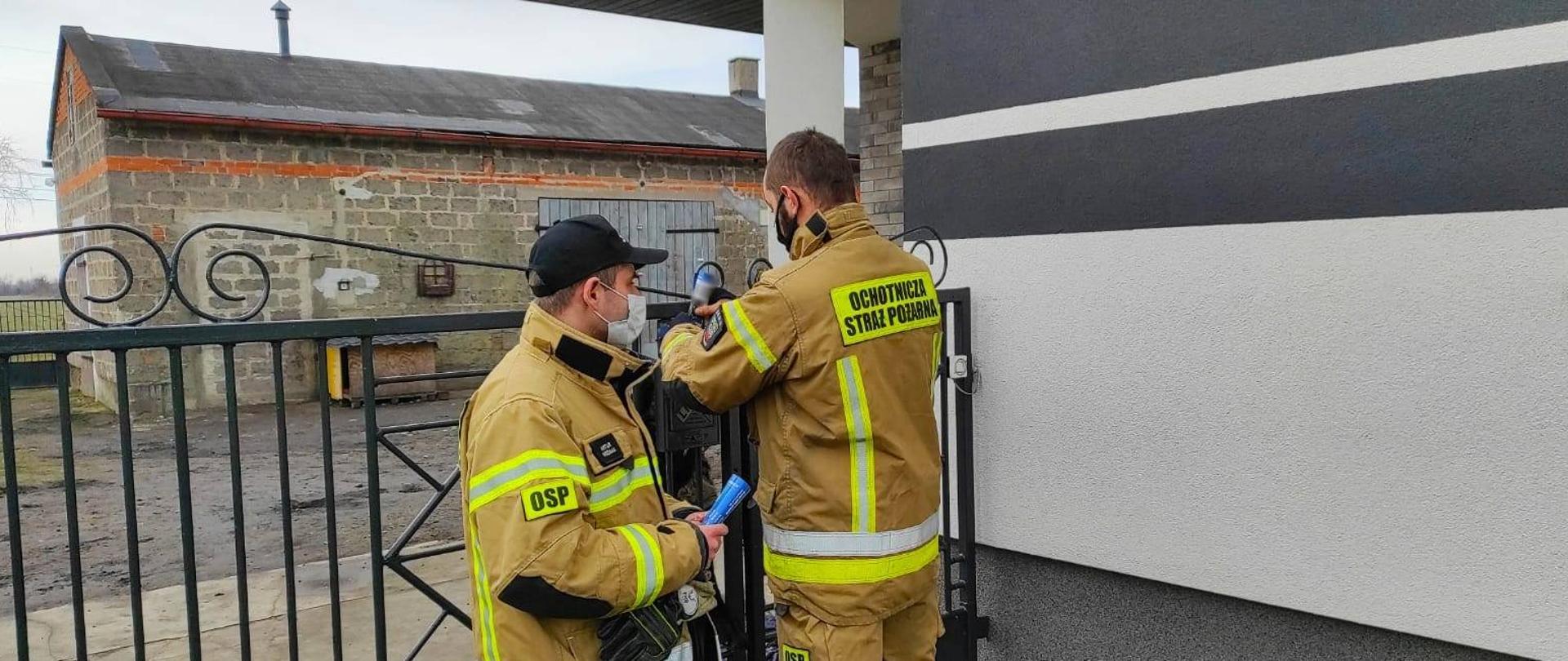 Dwóch strażaków OSP w mundurach typu Nomex roznoszą ulotki informacyjne dotyczące szczepień przeciw COVID-19. W tle posesja, brama koloru czarnego i budynki gospodarcze.