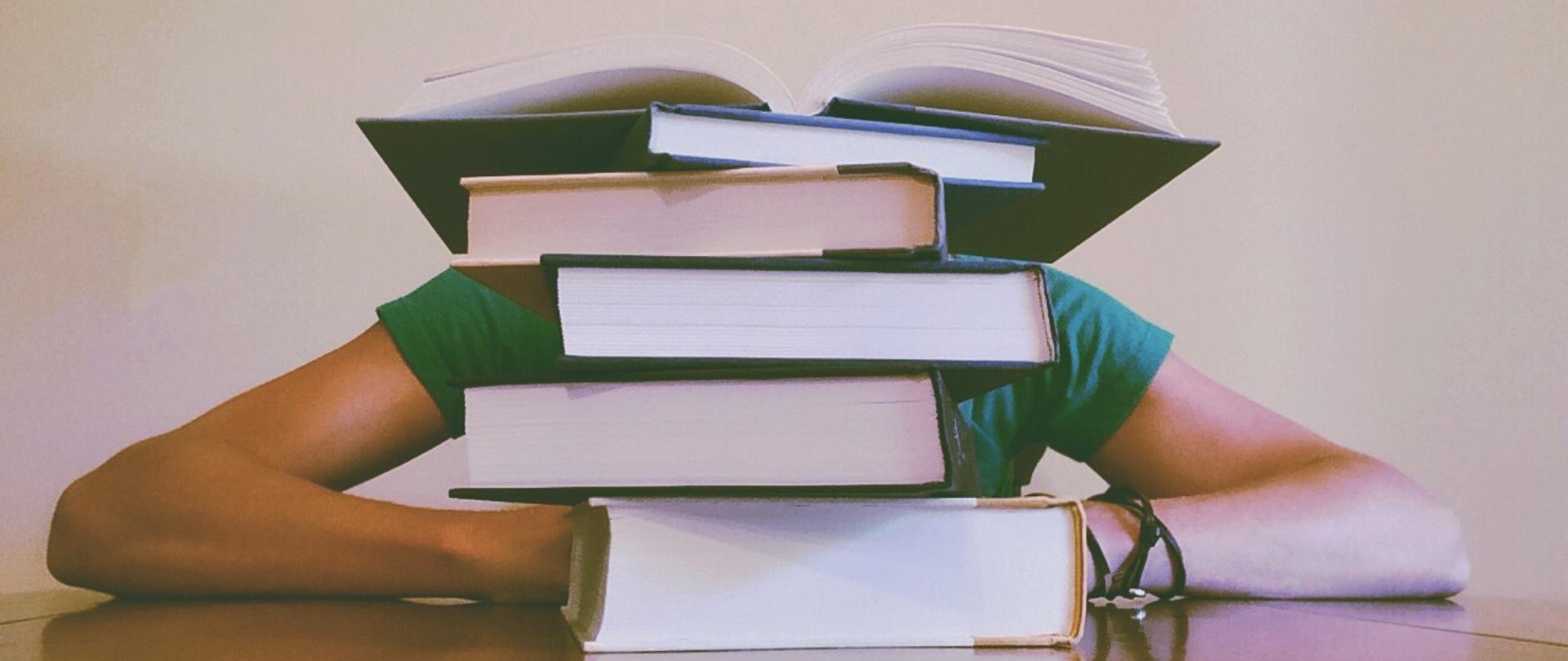 Zdjęcie przedstawiające książki ułożone jedna na drugiej na ławce szkolnej. Za książkami siedzi oparty o ławkę uczeń.