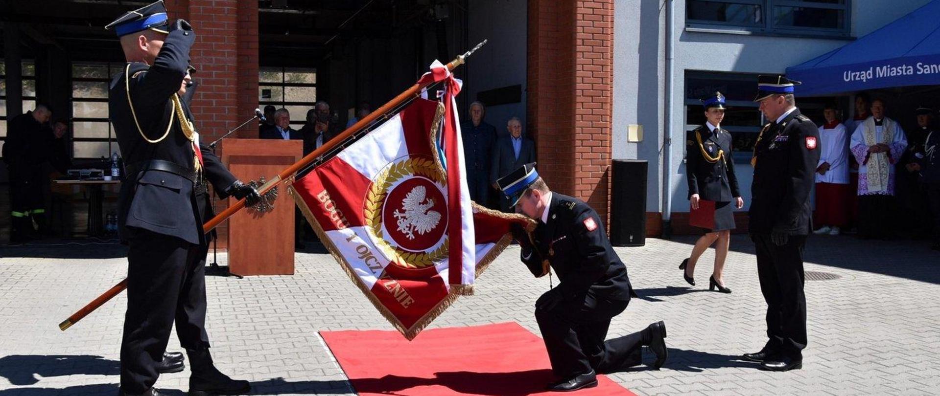 Zdjęcie zrobione na zewnątrz, podczas uroczystych obchodów Dnia Strażaka. Zdjęcie zrobione na placu przed jednostką ratowniczo-gaśniczą. Po lewej stronie stoi poczet sztandarowy trzymając pochylony sztandar komendy. Przed pocztem klęczy oficer Państwowej Straży Pożarnej i całuje materiał sztandaru.