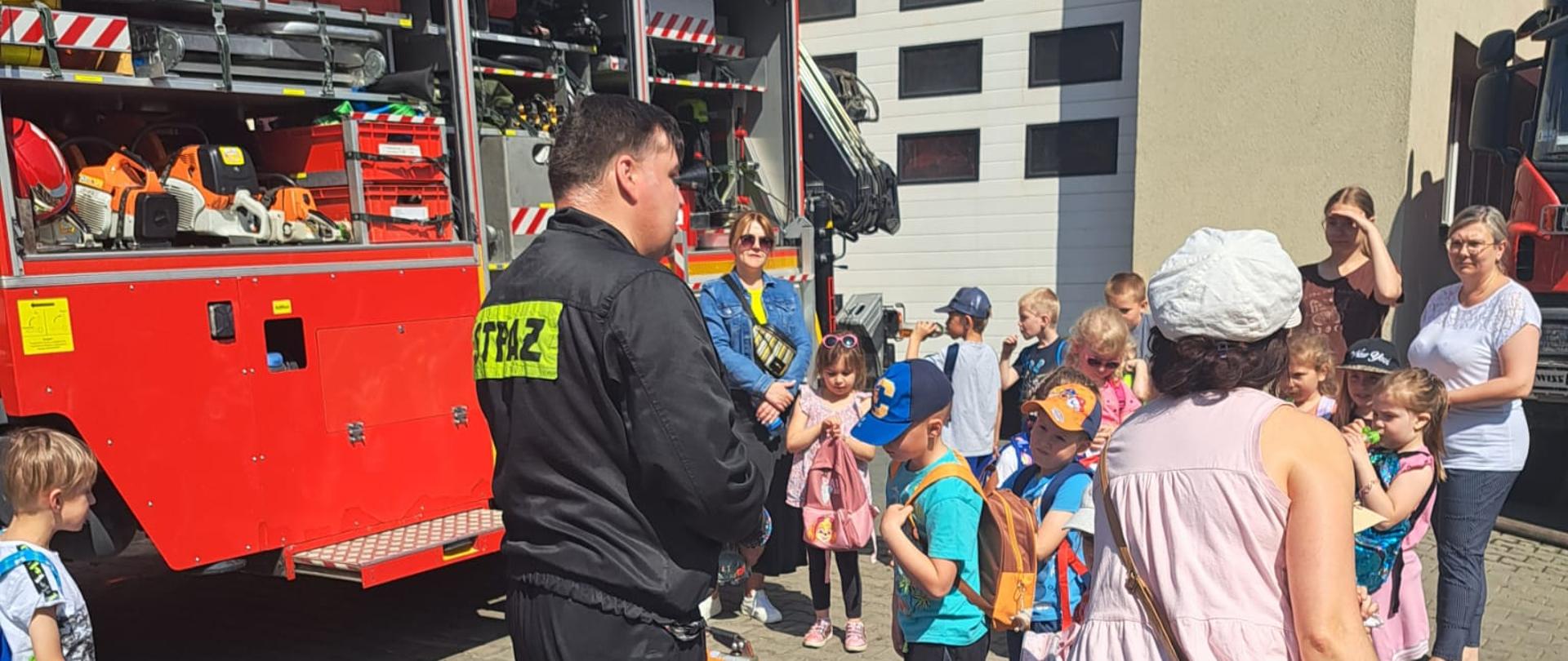 Dzieci i opiekunowie oglądają sprzęt i wóz strażacki na placu JGR. Obok strażak tłumaczący. 