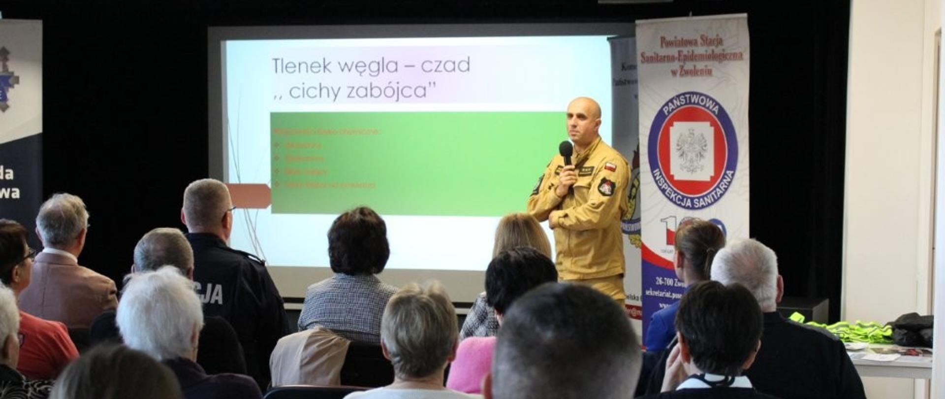 Na pierwszym planie widać tyły głów i ramiona osób siedzących na krzesłach, tyłem do kamery, oglądających prezentację. Na podium stoi łysy mężczyzna w beżowym mundurze, trzymający mikrofon i patrzący w lewo. Za nim znajduje się duży ekran, na którym wyświetlana jest prezentacja z tytułem w języku polskim: "Tlenek węgla – czad. „Cichy zabójca”". Po prawej stronie podium widoczny jest pionowy baner z logo Państwowej Inspekcji Sanitarnej w Zwoleń (Państwowa Stacja Sanitarno-Epidemiologiczna w Zwoleń). Oświetlenie jest skupione na prelegencie i ekranie, podczas gdy publiczność jest lekko zacieniona.