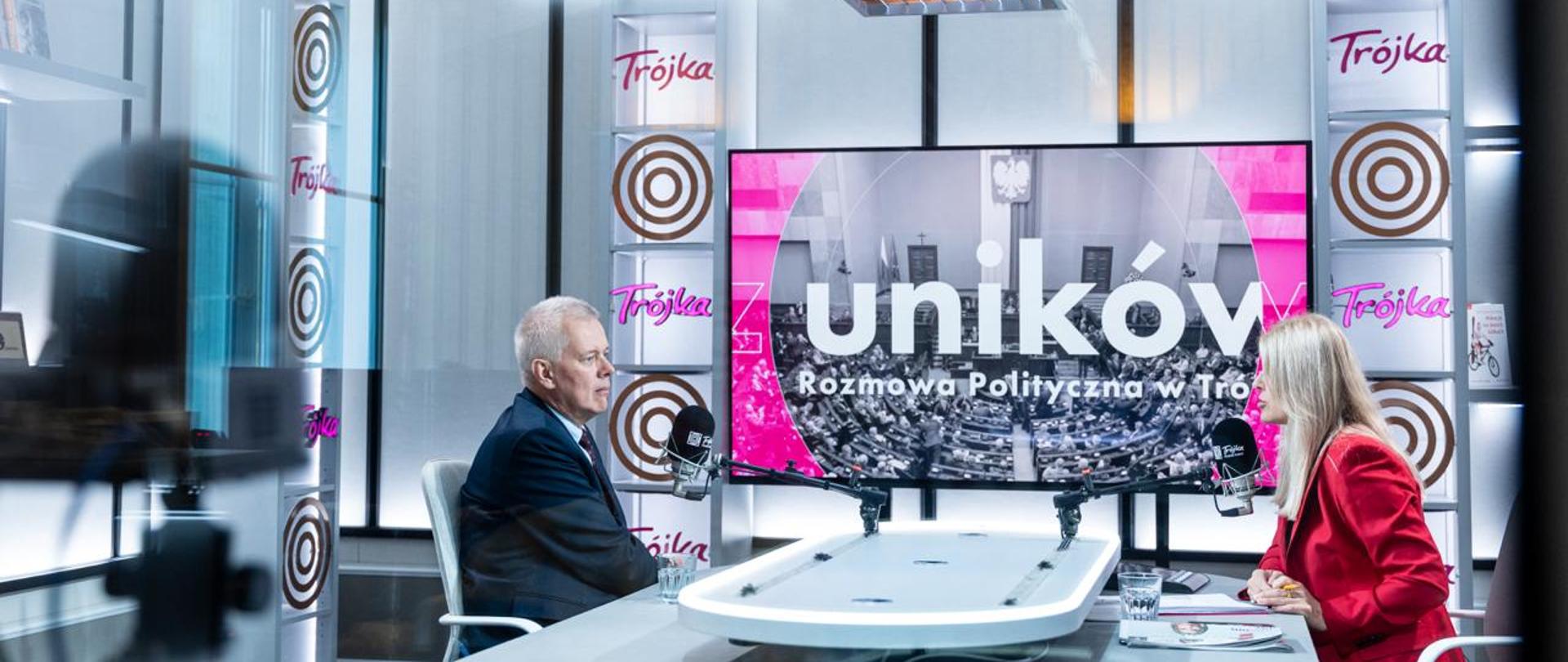 Na zdjęciu studio radiowe. Przy białym stole za mikrofonami siedzą: minister Tomasz Siemoniak (po lewej) oraz redaktor Renata Grochal (po prawej). Pomiędzy ministrem i prowadzącą telewizor z grafiką, przedstawiającą zdjęcie sali plenarnej Sejmu z napisem „Bez Uników, Rozmowa Polityczna w Trójce”. W tle widać różowe neony "Trójka" oraz inne elementy dekoracji studia. U góry zdjęcia oświetlenie studia.
