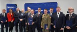 Zdjęcie zbiorowe, na tle granatowej ścianki z napisem NCBR stoi grupa dwunastu osób, niektórzy trzymają granatowe teczki.
