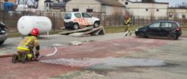 a zdjęciu przedstawiono ćwiczenia strażackie na otwartym placu. Dwóch strażaków w pełnym umundurowaniu ochronnym pracuje z linią gaśniczą – jeden z nich klęczy, podłączając wąż do rozdzielacza, natomiast drugi kieruje się w stronę pojazdów, trzymając prądnicę wody.
W centrum sceny znajduje się biały samochód ustawiony na specjalnej platformie treningowej, przeznaczonej do symulacji akcji ratowniczych. Obok widoczny jest czarny samochód osobowy. W tle znajdują się zabudowania oraz zbiornik, który może służyć do przechowywania gazu lub innych substancji.
Całość przedstawia realistyczny scenariusz szkoleniowy, mający na celu doskonalenie umiejętności strażaków w zakresie działań ratowniczo-gaśniczych oraz pracy z wyposażeniem.