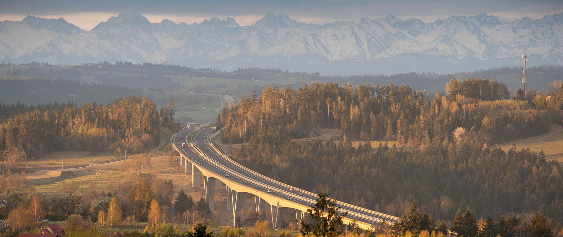 estakada w górzystym terenie, dookoła lasy, pola, nieliczne zabudowania, w tle wysokie góry