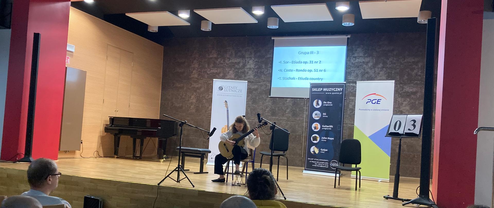 Uczennica naszej szkoły Amelia Lewkowicz podczas występu na XX Karnawałowym Konkursie Gitarowym "Z gitarą w Nowy Rok - Ok!" sali koncertowej w Państwowej Szkole Muzycznej I stopnia im. Zygmunta Noskowskiego w Skarżysku – Kamiennej w dniu 27 stycznia 2023 r. Uczennica ubrana na galowo - biała bluzka i czarne spodnie, siedzi na środku dużej sceny na krześle trzymając gitarę. Przed nią stoją dwa mikrofony służące do nagłośnienia występu. W tle na scenie stoją trzy banery sponsorów konkursu - jeden baner - biały z gitarą klasyczną - gitary lutnicze, drugi koloru niebieskiego - sklep muzyczny www. Quena.pl. trzeci baner z logo PGE. Na ścianie na rzutniku wyświetlony jest program, który wykonuje uczennica - Grupa III - 3 oraz program:
F. Sor - Etiuda op. 31 nr 2
N. Coste. Rondo op. 51 nr 6
T. Stachak - Etiuda country
Na dole zdjęcia widać od tyłu siedzenia widowni sali koncertowej w Skarżysku - Kamiennej i ludzi siedzących na nich.