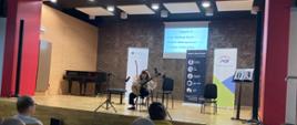 Uczennica naszej szkoły Amelia Lewkowicz podczas występu na XX Karnawałowym Konkursie Gitarowym "Z gitarą w Nowy Rok - Ok!" sali koncertowej w Państwowej Szkole Muzycznej I stopnia im. Zygmunta Noskowskiego w Skarżysku – Kamiennej w dniu 27 stycznia 2023 r. Uczennica ubrana na galowo - biała bluzka i czarne spodnie, siedzi na środku dużej sceny na krześle trzymając gitarę. Przed nią stoją dwa mikrofony służące do nagłośnienia występu. W tle na scenie stoją trzy banery sponsorów konkursu - jeden baner - biały z gitarą klasyczną - gitary lutnicze, drugi koloru niebieskiego - sklep muzyczny www. Quena.pl. trzeci baner z logo PGE. Na ścianie na rzutniku wyświetlony jest program, który wykonuje uczennica - Grupa III - 3 oraz program:
F. Sor - Etiuda op. 31 nr 2
N. Coste. Rondo op. 51 nr 6
T. Stachak - Etiuda country
Na dole zdjęcia widać od tyłu siedzenia widowni sali koncertowej w Skarżysku - Kamiennej i ludzi siedzących na nich.