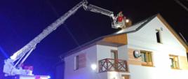 Zdjęcie przedstawia budynek mieszkalny, parterowy. Przed nim znajduje się samochód specjalny z rozłożonym parkiem drabinowym. W jego koszu znajdują się strażacy w umundurowaniu specjalnym z czerwonymi hełmami na głowach. Strażacy pracują przy obtrącaniu sadzy w przewodzie kominowym. Teren działań oświetlony- zdjęcie zrobione w porze nocnej. 