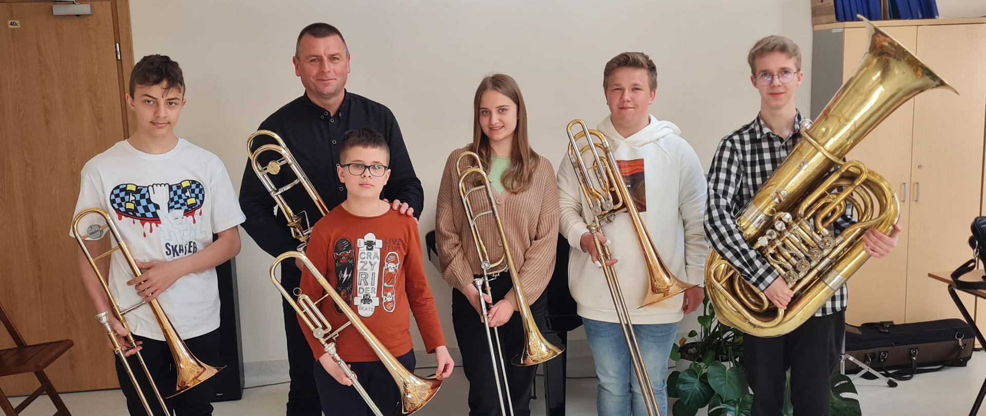 Zdjęcie. Grupa uczniów i nauczyciel prowadzący podczas warsztatów puzonowych . Wszyscy trzymają w rękach swoje instrumenty - puzony. Tylko jeden uczeń - ostatni z prawej strony trzyma tubę.