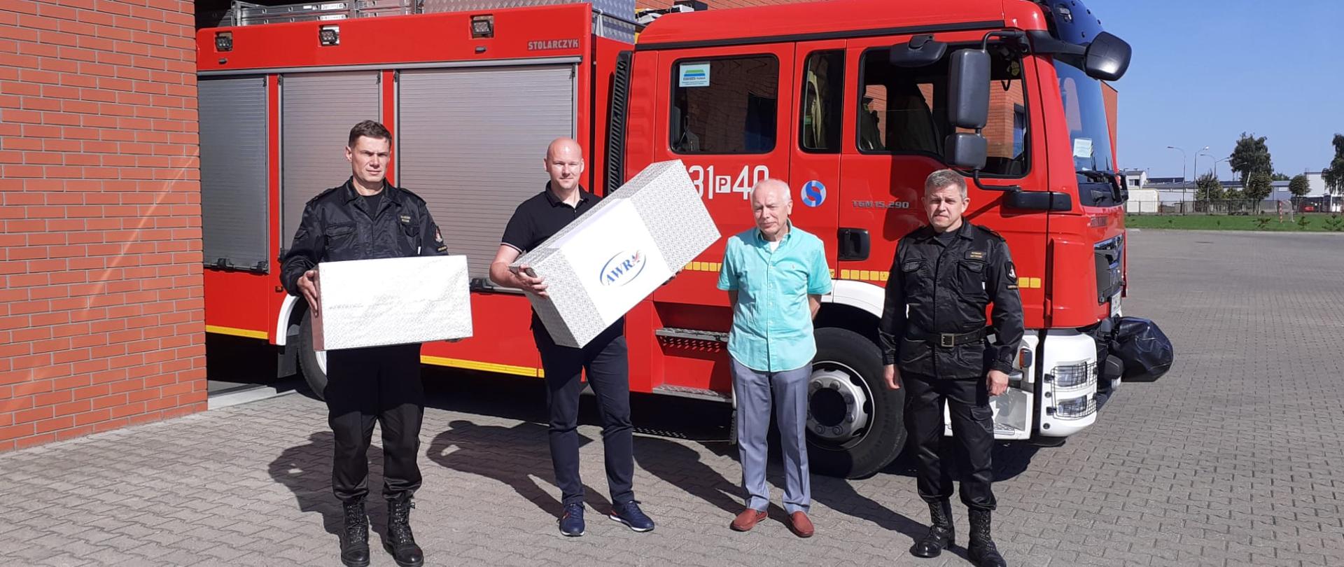 Dwóch funkcjonariuszy i dwóch przedstawicieli firmy AWRA stoi na tle samochodu pożarniczego, trzymając dwa kartony.