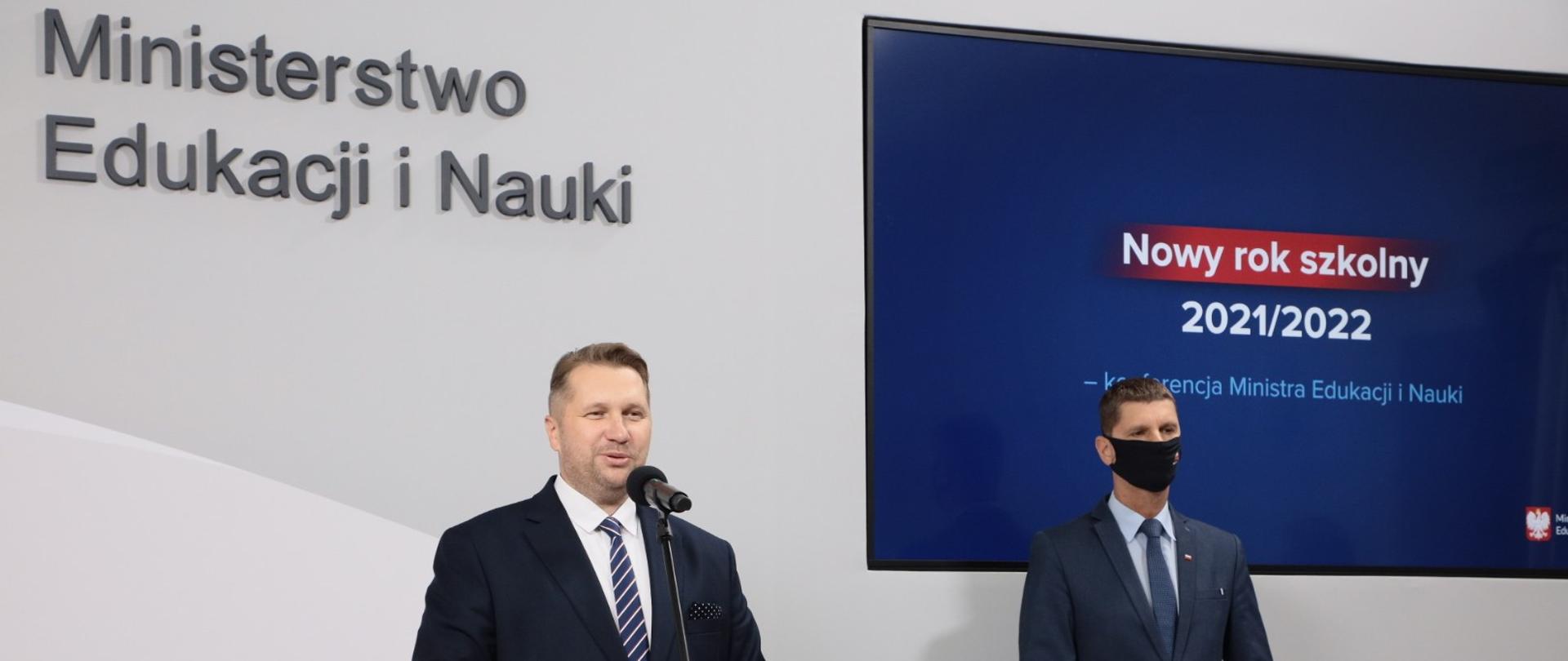 Minister Przemysław Czarnek i wiceminister Dariusz Piontkowski przemawiają podczas konferencji dotyczącej nowych zasad w roku szkolnym 2021/2022. W tle napis Ministerstwo Edukacji i Nauki. Po prawej stronie na telewizorze wyświetlona prezentacja. 