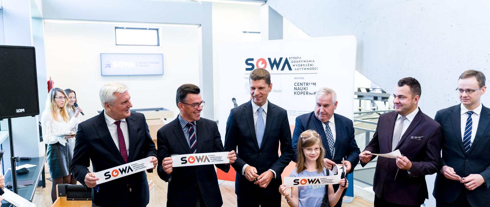 Sześciu mężczyzn i dziewczynka trzymają fragmenty przeciętej wstęgi z napisem SOWA 