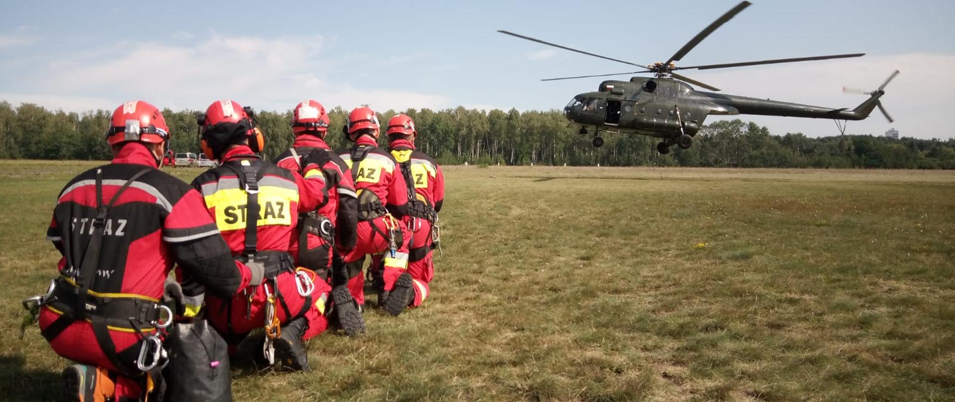 5 strażaków w czerwonych kombinezonach w pozycji klęczącej na jednym kolanie na trawie oczekuje na lądowanie policyjnego śmigłowca