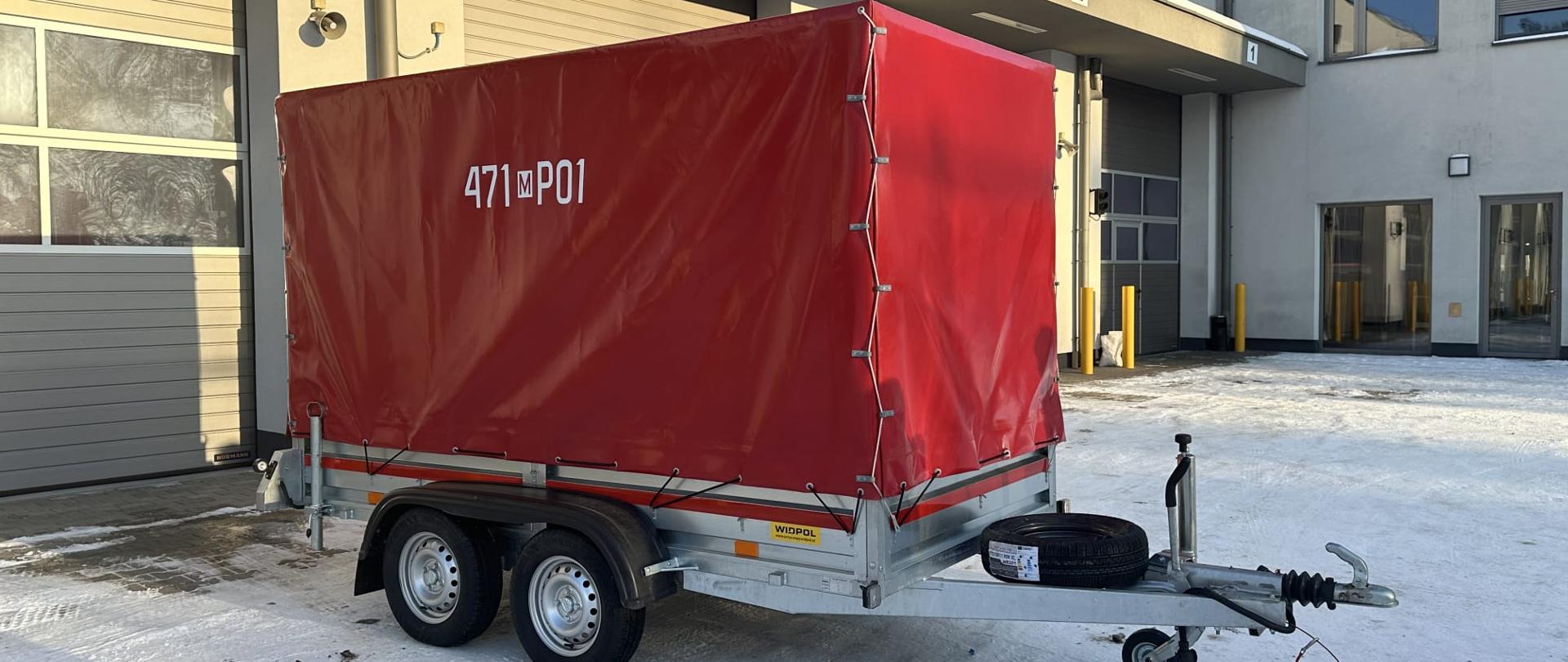 Na zdjęciu widać dwuosiową przyczepę transportową należącą do straży pożarnej, zaparkowaną przed budynkiem remizy. Przyczepa ma metalową ramę i jest przykryta czerwoną plandeką, na której widnieje oznaczenie operacyjne „471[M]P01”.
Z przodu znajduje się dyszel z zaczepem do podłączenia do pojazdu oraz koło podporowe. Na ramie zamocowane jest także koło zapasowe. Przyczepa stoi na dwóch osiach z czterema kołami, co sugeruje, że jest przeznaczona do przewożenia cięższego sprzętu.
W tle widoczny jest budynek straży z kilkoma bramami garażowymi oznaczonymi numerami. Jedna z bram jest przeszklona, inne są zamknięte. Na ziemi widać śnieg i lód, co wskazuje na zimowe warunki, natomiast słońce świeci jasno, rzucając wyraźne cienie.