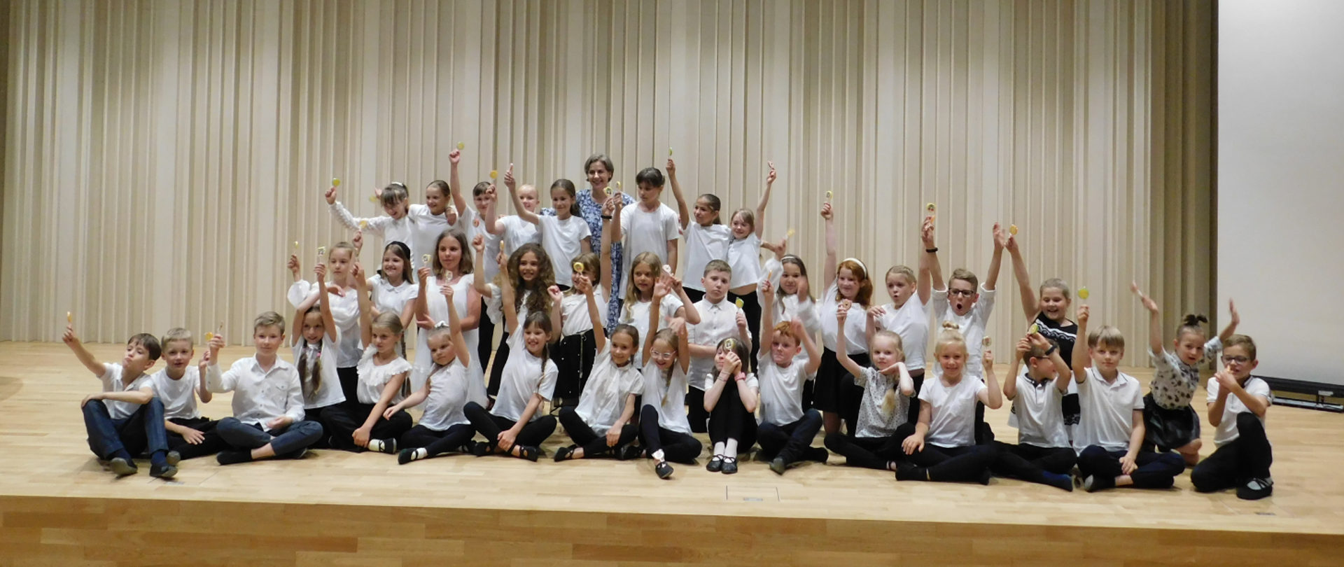 Na zdjęciu duża ilość dzieci, wszyscy ubrani w białe koszulki i czarne spodnia. Każdy trzyma lizaka.