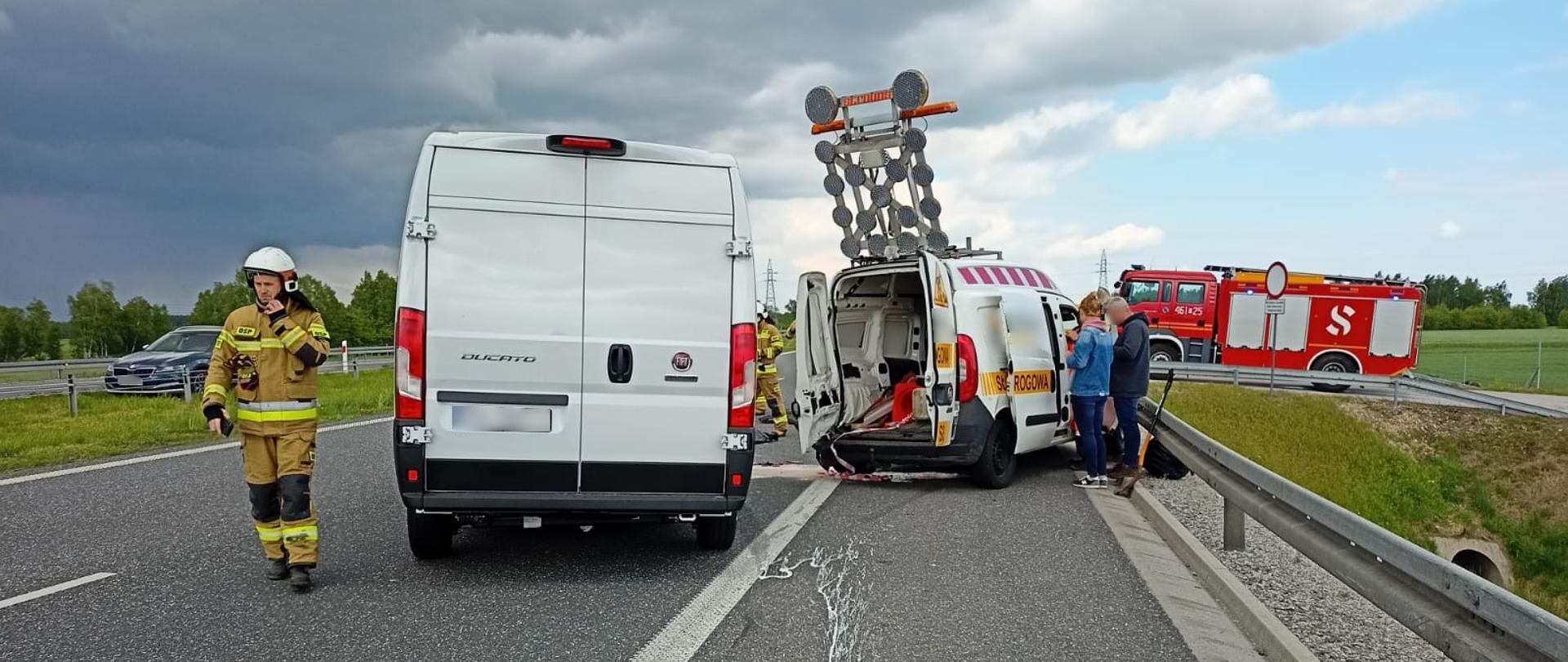 Miejsce zdarzenia widoczne samochodu typu bus uszkodzone w wyniku zderzenia oraz służby ratownicze/ strażacy działający na miejscu.