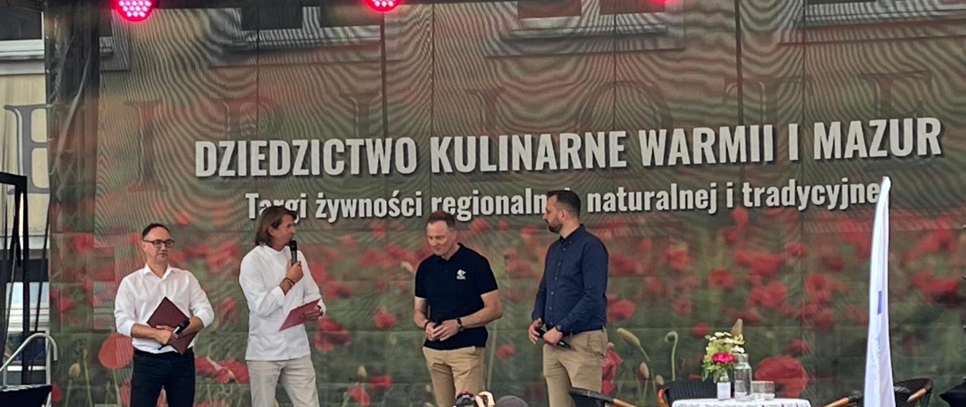 Na zdjęciu znajduje się prowadzący wydarzenie Targi żywności regionalnej, naturalnej i tradycyjnej. Dziedzictwo Kulinarne Warmii i Mazur, gość specjalny wydarzenia Karol Okrasa, Marszałek Województwa Warmińsko – Mazurskiego oraz Prezydent Olsztyna