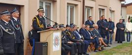 zastępca komendanta głównego psp nadbrygadier Krzysztof Hejduk przemawia przy mównicy, z lewej dwóch druhów w mundurach galowych po prawej strażacy psp i osp w mundurach galowych oraz ludzie w odświętnych ubraniach za nimi budynek