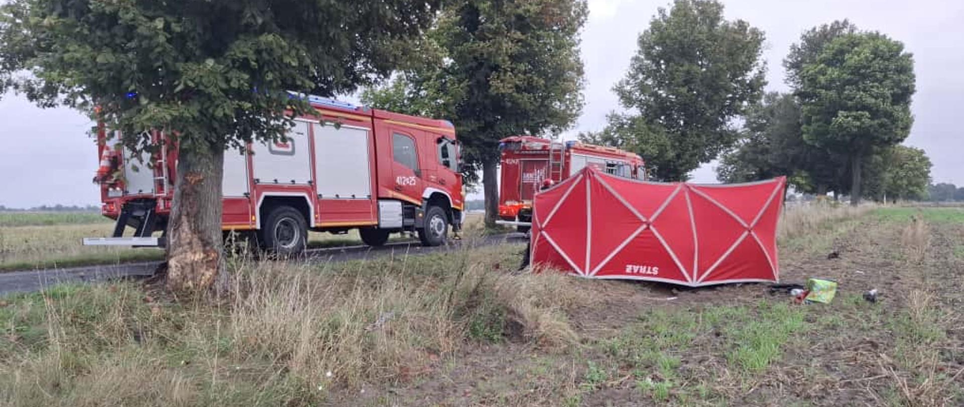 Rozległy kadr ukazujący pojazdy służb ratowniczych oraz fragment miejsca wypadku zabezpieczonego przez strażaków.