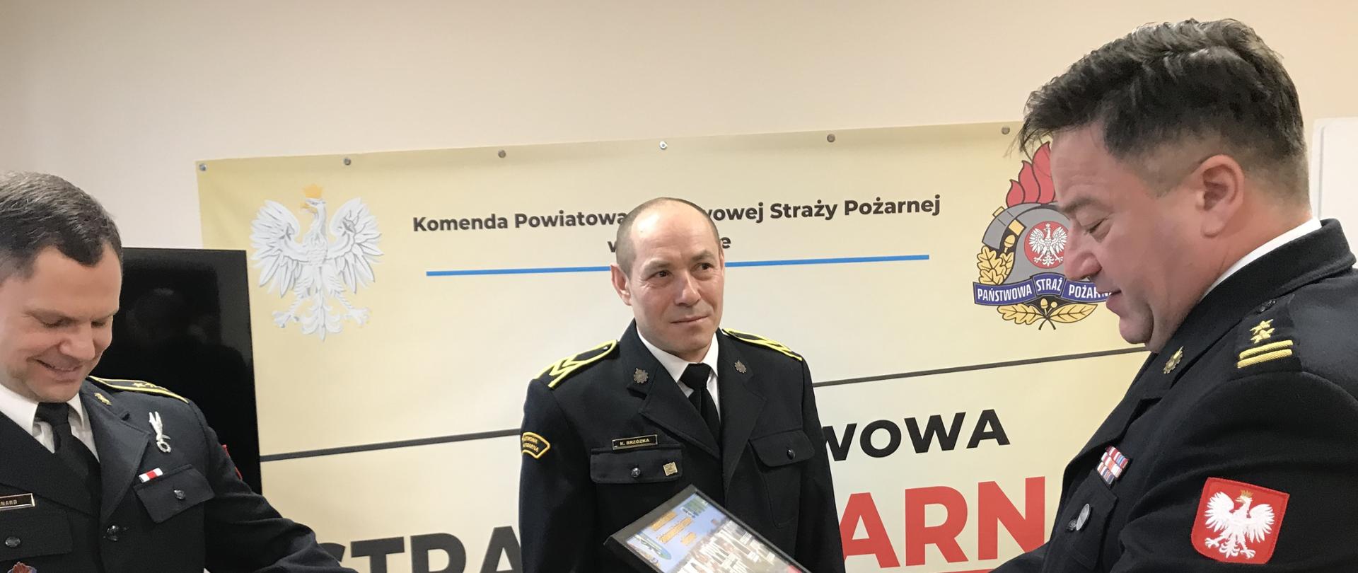 Zdjęcie zrobione na świetlicy Komendy Powiatowej PSP. Funkcjonariusze ubrani w czarny mundur wyjściowy. Komendant Powiatowy trzyma w ręku kolorowe tablo ze zdjęciami funkcjonariuszy i pracowników komendy. Od lewej strażak który otrzymał tablo, po środku strażak oczekujący na wręczenie i po prawej Komendant wręczający tablo. W tle ścianka koloru piaskowego.