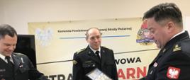 Zdjęcie zrobione na świetlicy Komendy Powiatowej PSP. Funkcjonariusze ubrani w czarny mundur wyjściowy. Komendant Powiatowy trzyma w ręku kolorowe tablo ze zdjęciami funkcjonariuszy i pracowników komendy. Od lewej strażak który otrzymał tablo, po środku strażak oczekujący na wręczenie i po prawej Komendant wręczający tablo. W tle ścianka koloru piaskowego.