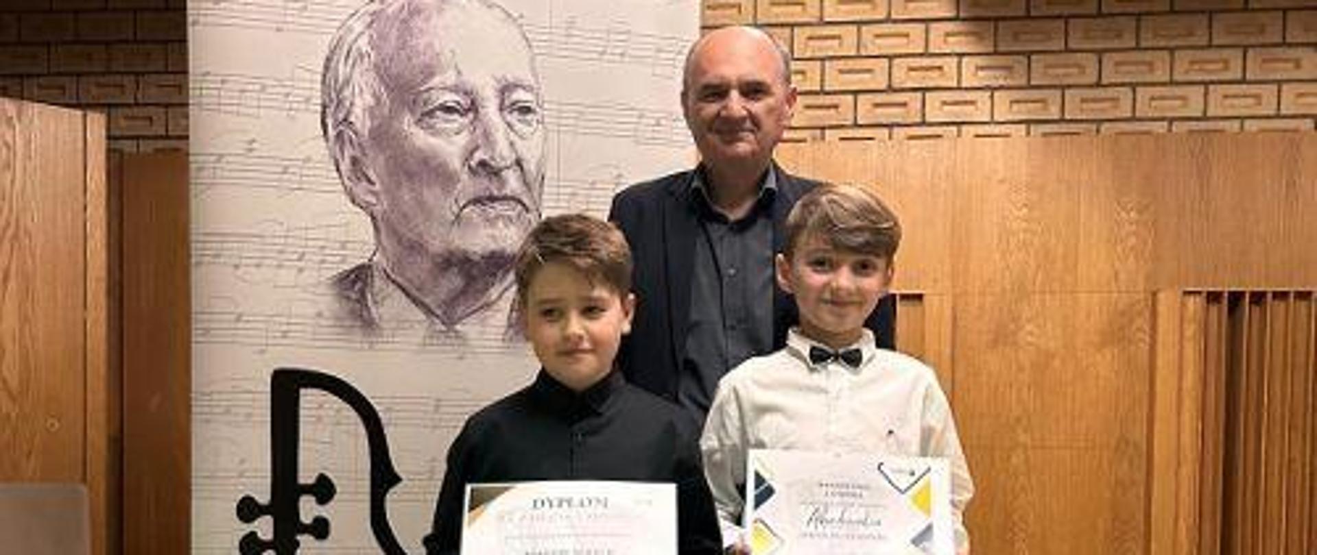 Dwóch uczniów trzymających dyplomy laureatów wraz z nauczycielem po środku.