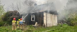 Budynek parterowy, którego dach znajduje się w lekkim zadymieniu. Przed domem trzech strażaków w umundurowaniu specjalnym, którzy podają jeden prąd wody w natarciu na palący się dach.