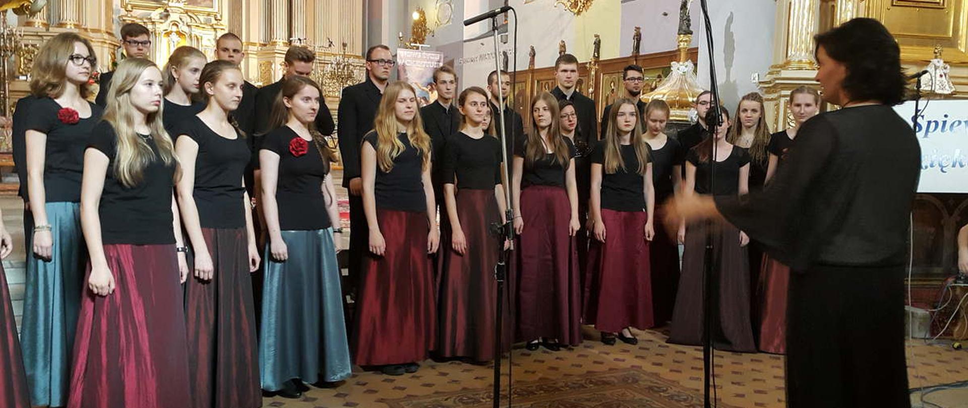 Kolorowe zdjęcie z występu chóru, chórzyści stoją w dwóch, półokrągłych rzędach, dziewczyny ubrane są w czarne koszulki i długie bordowe lub zielone spódnice, chłopacy w czarne koszule i ciemne garnitury