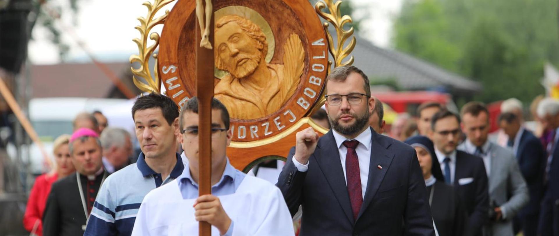 Minister Rolnictwa i Rozwoju Wsi Grzegorz Puda niesie obraz religijny Świętego Andrzeja Boboli w ozdobnych ramach lub posąg umieszczony na piedestale, do którego przymocowane są drążki służące noszeniu w czasie procesji.