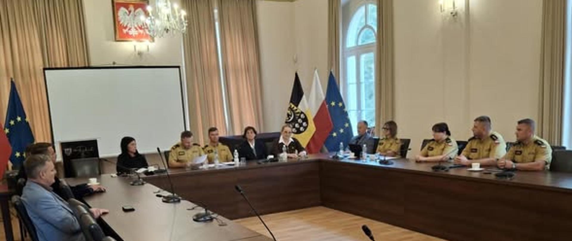 Na zdjęciu widać spotkanie przy dużym, ustawionym w kształcie litery „U” stole konferencyjnym. Przy stole siedzą przedstawiciele Państwowej Straży Pożarnej oraz zaproszeni goście. W tle widoczne są polskie i unijne flagi oraz symbole strażackie. To spotkanie założycielskie Społecznego Komitetu Fundacji Sztandaru dla Komendy Powiatowej Państwowej Straży Pożarnej.