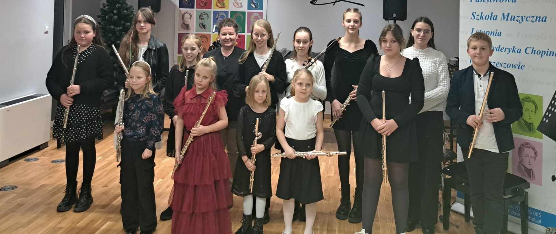 Uczniowie wraz z pedagoggiem pozują do zdjęcia stojąc w dwurzędzie i trzymając w dłoniach flety. W tle po prawej widać baner szkoły muzycznej w Ostrzeszowie, po lewej choinkę a z tyłu po obu stronach kolumny na statywach i kolorową grafikę przedstawiającą F. Chopina.