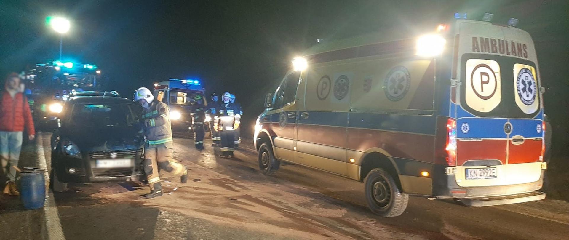 Samochód uczestniczący w zdarzeniu oraz karetka Pogotowia Ratunkowego. Obok pojazdów strażacy oraz oraz osoby uczestniczące w zdarzeniu. 